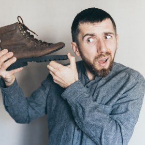 Young bearded Caucasian man holds old leather boot with torn sole, listening, surprise and shock on his face