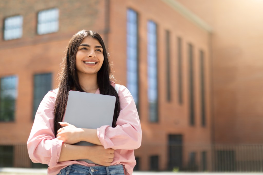 Cheerful young girl student holding a closed laptop, looking upwards thoughtfully beside a university building SSUCv3H4sIAAAAAAAACnSSTWvDMAyG74P9B+NzCk3StEuvY/fBtlPZQZHVxNS1g+10lNL/PiduOg82yCF6JL368uXxgTHegJPIt+wyWsGWSg3OW/DS6ICX2Y1b0oJsSk62XC9TADK1SEhvrASVwgY8dhqOFKAelBrxdXJy58EPjtzYyw0heGqDRoR3idjwLtpsdkzOkBFc/JVMr4hniccNzeh5J9KOzzzW+UfkRQwY1/CHzht2xqiMPRulqKWMfWh5IuukPzPy+FMh/nzed9SSxvM0YzK7JUUQZ9/FUH748mSP6TZgENIkizgZBDUGlIkSyB4s/MrrrUSp24QY302nnJXQDNrbsSte5PU8LlfG9NCEPW7ZPlSimXfgXMgQM0/KY3g75piU0sZPY/GoykU46WjmZVlVdZVX1eapDl+xuQXEZ9DJoDN1NAsFGw9SpIPJsQNeIOWVyHGBRQGLVb0uFs1mj4tSNKt93gCKVR3Ocf0GAAD//wMAzR4OcO8CAAA=