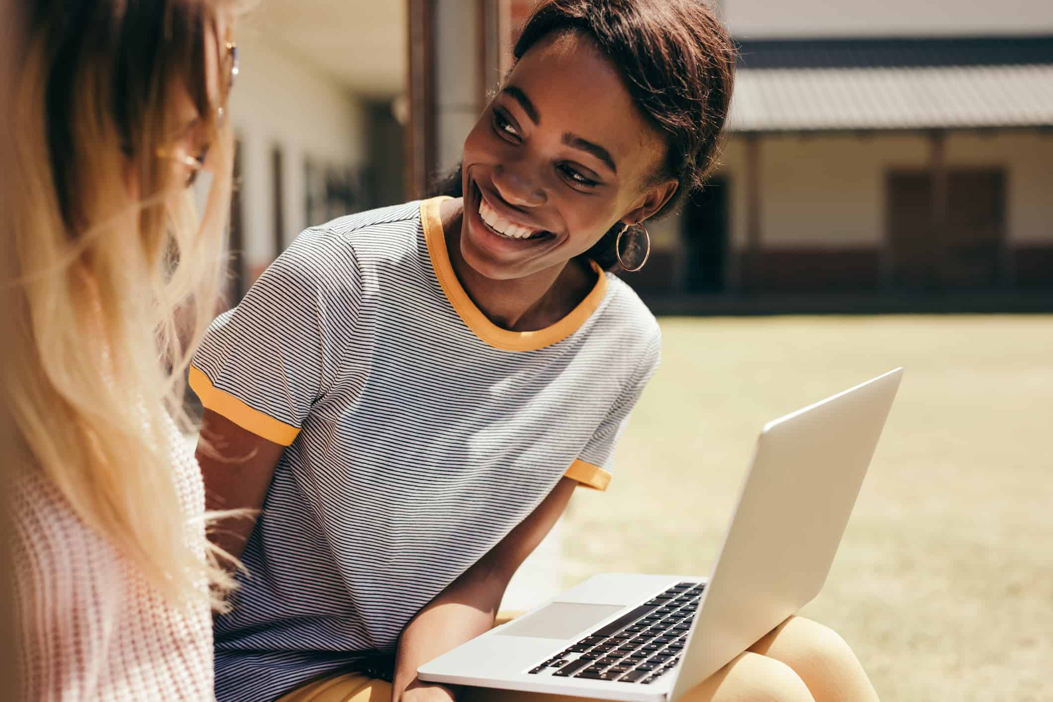 Smiling female students sitting at college campus with laptop computer. Two young students sitting at high school campus with laptop.